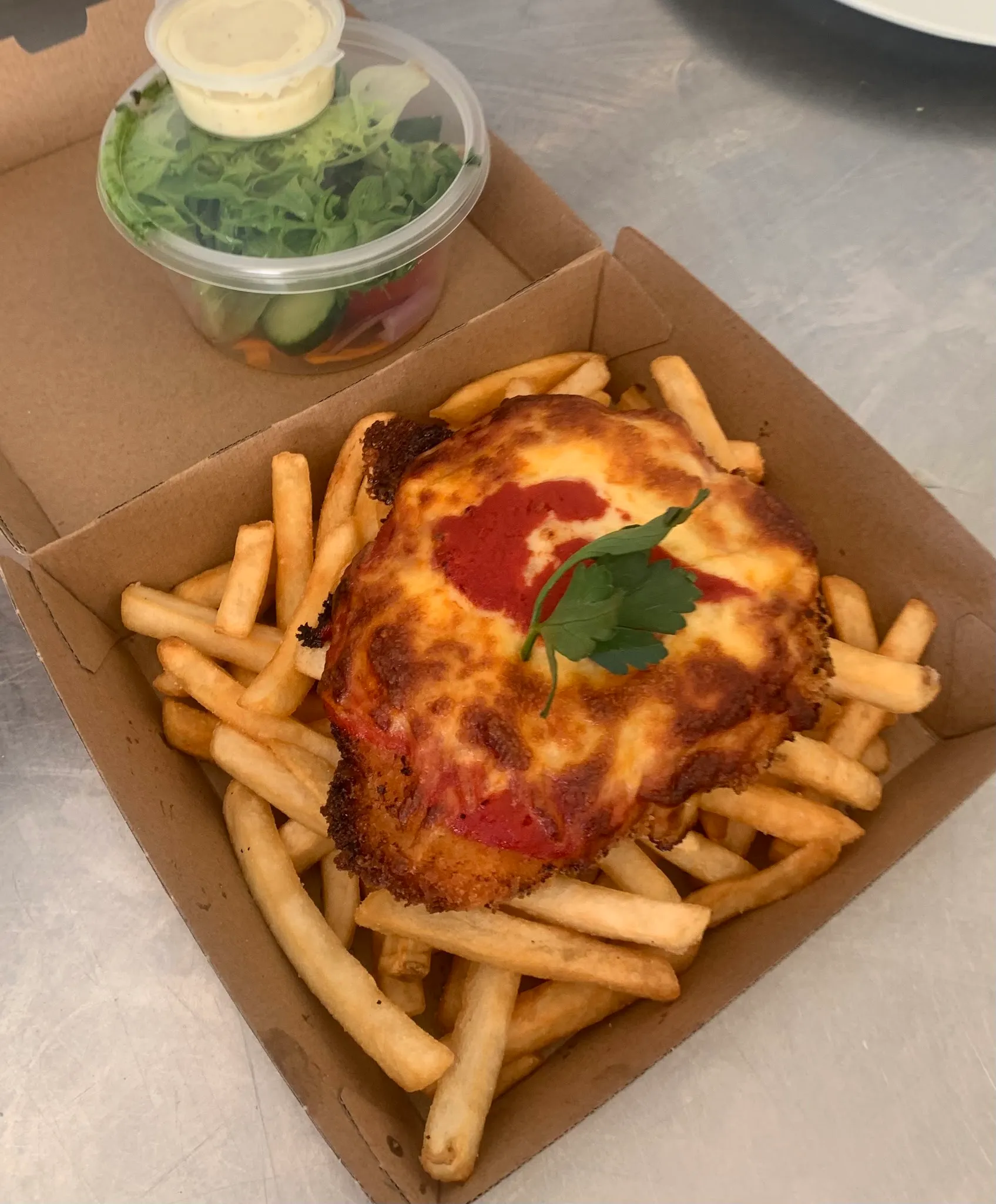 Chicken parmigiana and chips in a takeaway box with side salad